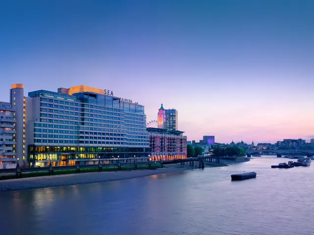 Dusk view of a large illuminated hotel building beside the River Thames, with boats on the water and the London Eye partly visible behind it.
