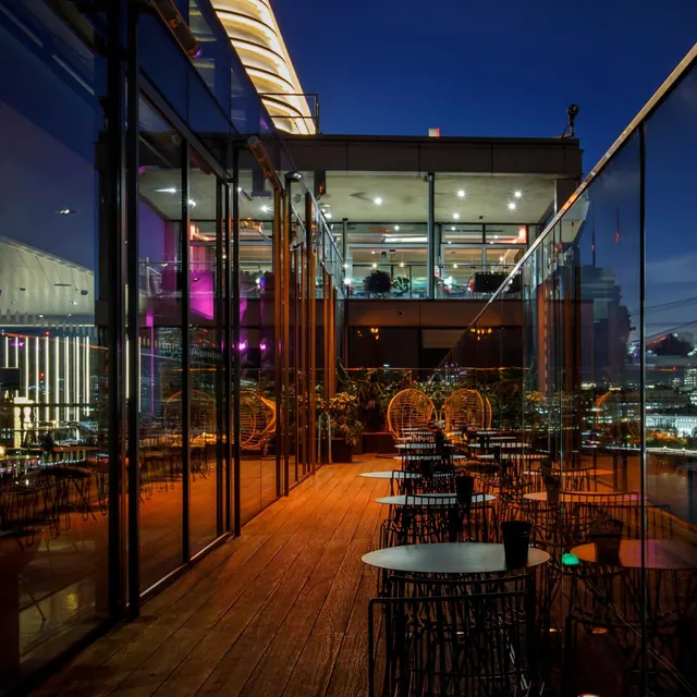 A rooftop terrace at dusk with wooden decking, black tables and chairs, glass walls and distant city lights.