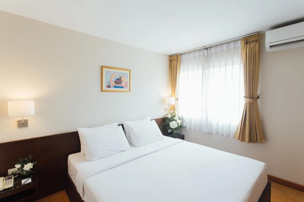 A made-up bed with white linens and pillows sits beside a window with gold curtains and a wall-mounted air conditioner.