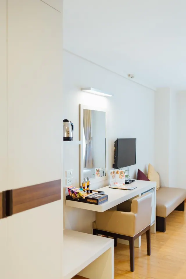 A hotel room with a white wall-mounted desk, mirror, flat-screen television, electric kettle, snack tray, and a beige bench seat over a wooden floor.