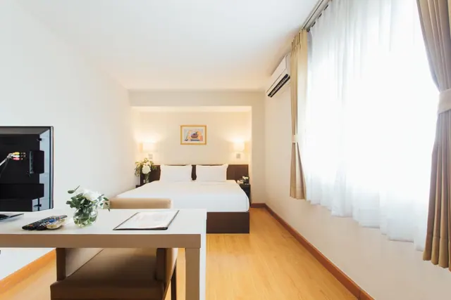 A hotel bedroom with a double bed against a white wall, a desk with a television and a flower vase in the foreground, and a wall-mounted air conditioner beside a window with sheer curtains.