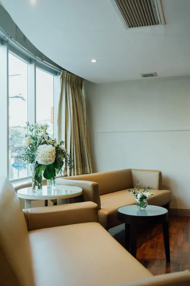 A lounge seating area with two tan leather sofas, round glass side tables holding white flower arrangements, and a curved window with cream curtains looking onto a street with trees.