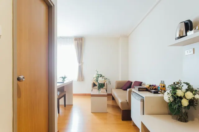 A hotel room living area with a beige sofa and burgundy cushions, a coffee table with a flower vase, a desk, a kettle on a wall shelf, and a minibar beside a vase of white flowers.
