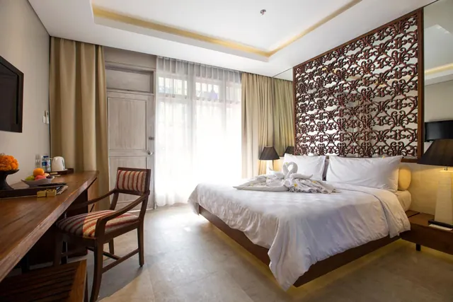 A bedroom with a king bed with towel swans on it, a wooden desk and chair, a wall-mounted television, and a window with sheer curtains.