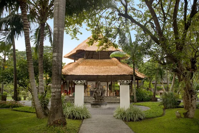 A thatched-roof Balinese pavilion with a stone statue stands at the end of a paved path through a green garden lined with palm trees.