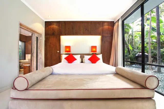 A king bed with red cushions and matching lamps sits against a carved wooden headboard wall, with floor-to-ceiling windows beside it looking onto a palm garden.
