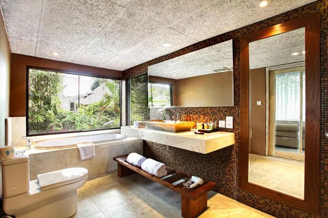 A bathroom with a built-in bathtub beside a window looking onto tropical plants, a long floating vanity with a basin and mirror, a toilet, and a wooden bench holding folded towels.