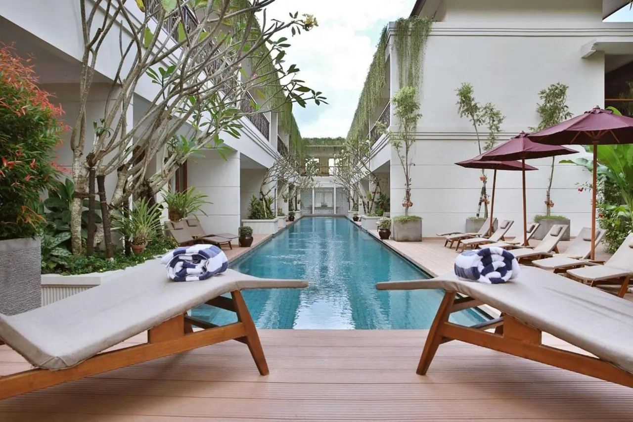 A long lap pool in a courtyard is flanked by wooden sun loungers with rolled towels and red parasols, with white building walls and trees around it.