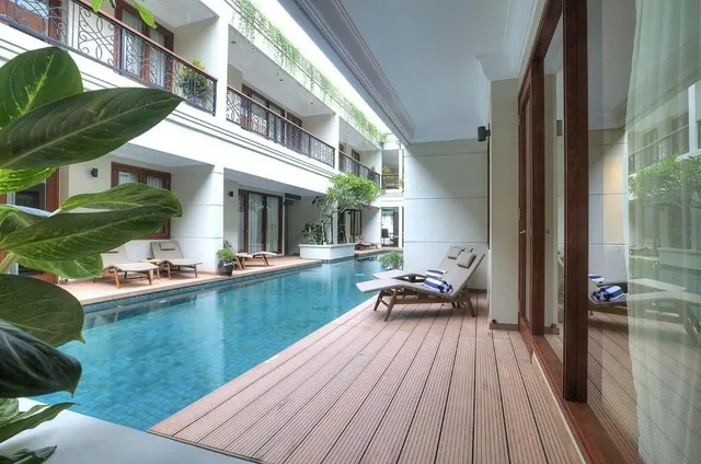 A long swimming pool with a timber deck, sun loungers with towels, and a multi-storey building with balconies around the courtyard.