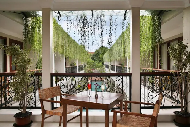 A covered balcony with a wooden table, two wicker chairs and two drinks, overlooking a courtyard with a pool and hanging greenery.