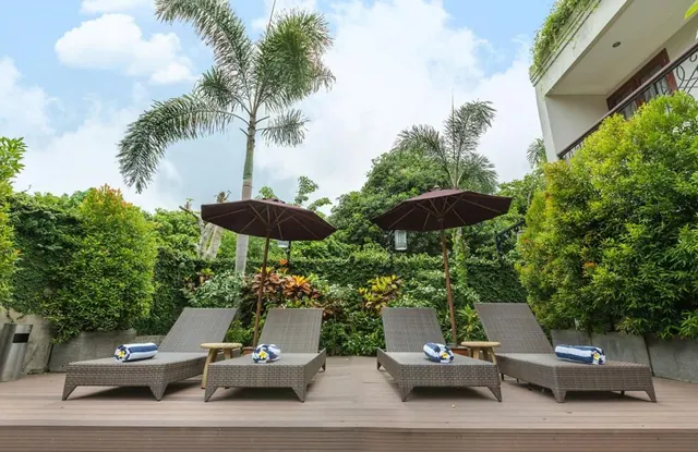 Four wicker sun loungers with rolled towels and two brown parasols stand on a timber deck in front of a green garden and a palm tree.