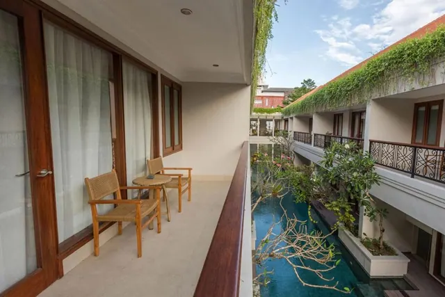 A covered balcony with two woven chairs and a small round table overlooks a narrow pool lined with frangipani trees.