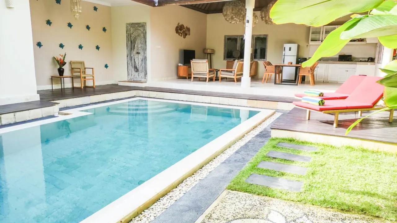A rectangular swimming pool sits beside a covered open-plan living area with armchairs, a dining table and a kitchenette, with red sun loungers on the deck at right.