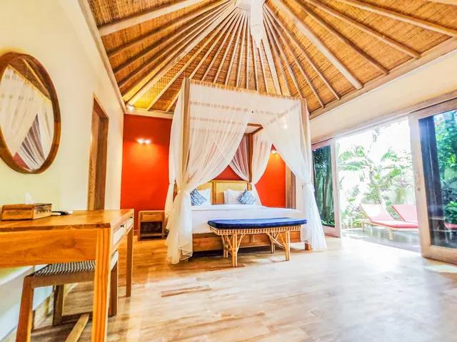 A bedroom with a canopy bed under a thatched ceiling, a wooden desk, and glass doors opening onto a terrace with red sun loungers and tropical plants.