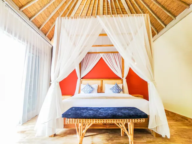 A bedroom with a canopy bed under a thatched ceiling, a red accent wall, a blue velvet bench at the foot of the bed, and sheer curtains beside a bright window.