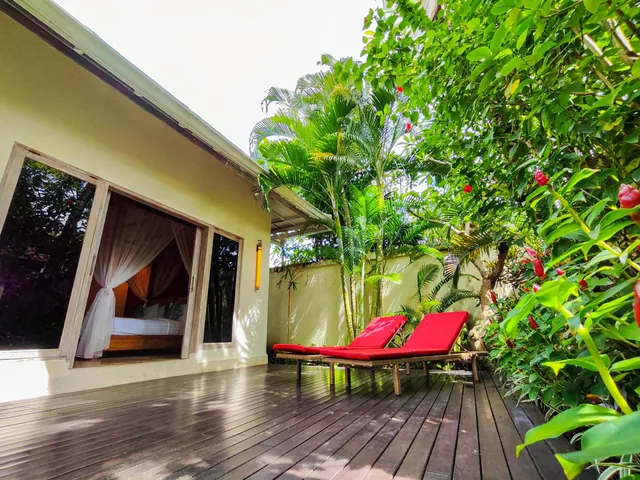 A wooden deck terrace with two red sun loungers sits beside an open sliding door to a bedroom with a bed under a mosquito net, facing a lush tropical garden.