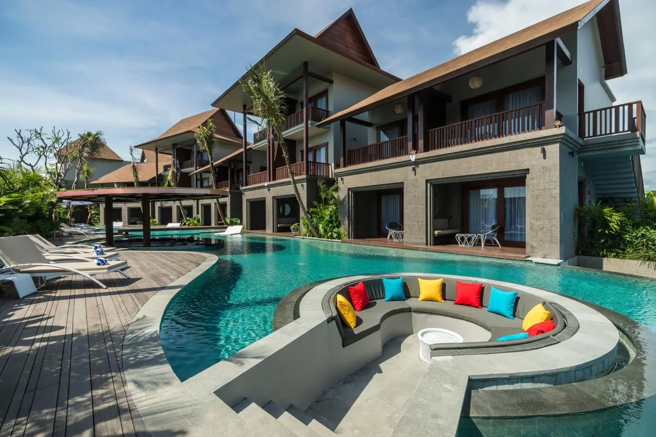 A turquoise pool with a curved, colourful cushioned seating area in the water, sun loungers on a timber deck, and a multi-storey Balinese-style building with balconies behind.