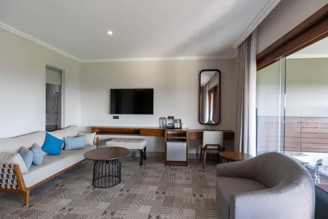 A hotel room living area with a cream sofa, wall-mounted television, a wooden desk with a kettle tray and small fridge, and a glass door leading to a balcony.