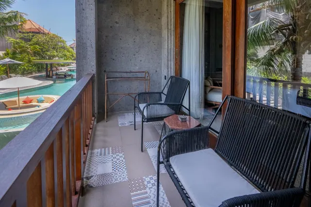 A private balcony with black woven chairs and a loveseat overlooks a swimming pool with sun loungers, umbrellas and tropical greenery.