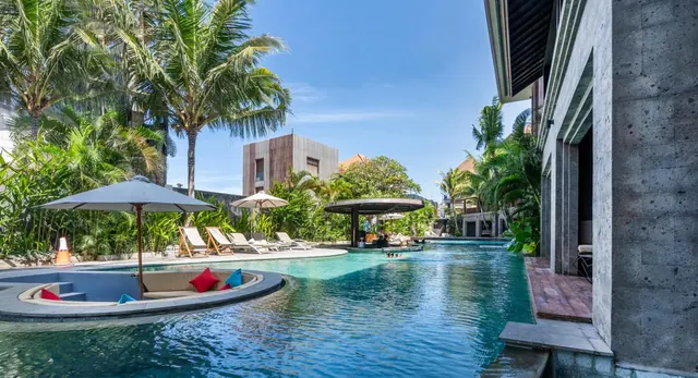 A long pool runs past sun loungers and shaded umbrellas, with palm trees, a sunken seating area and a concrete building beside it.