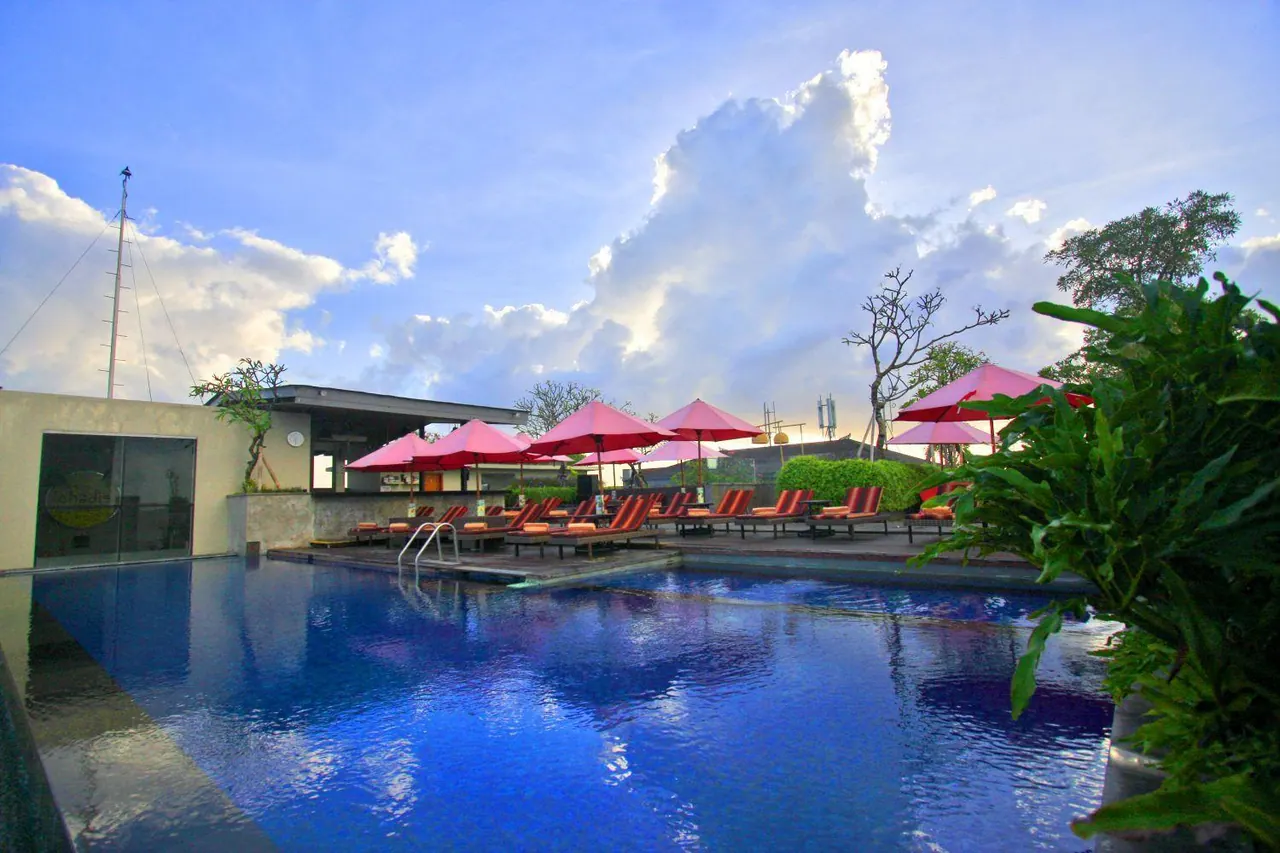 A large swimming pool with red umbrellas and sun loungers beside it under a partly cloudy sky.
