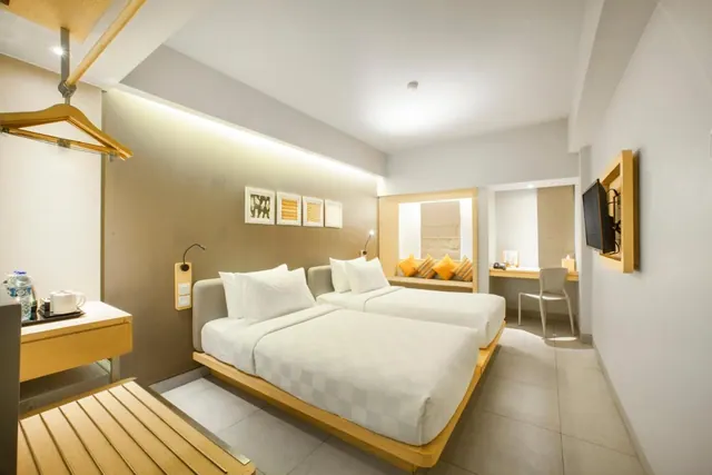 A bedroom with two white beds on a wooden platform, a wall-mounted television, a window seat with yellow cushions, and a work desk with a chair.
