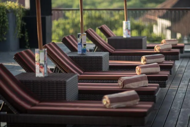 Rows of cushioned sun loungers with rolled towels and drink cartons on side tables sit on a wooden deck, with a railing, greenery and rooftops beyond.