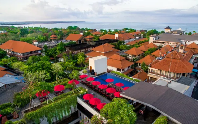 Aerial view of a resort pool with red umbrellas and sun loungers, surrounded by orange-roofed buildings and trees, with the ocean beyond.
