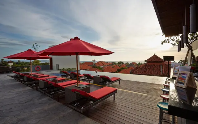 Red-cushioned sun loungers and red umbrellas line a pool deck beside an infinity pool, with red-tiled rooftops and the sea visible beyond.