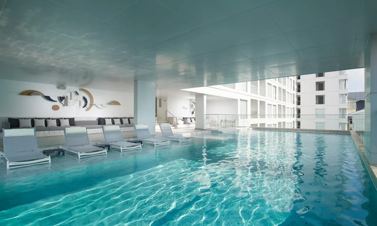 A covered pool with grey sun loungers and a sofa along the left wall, with a neighbouring building visible through the open side.