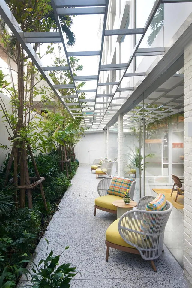 A covered walkway with a glass roof runs past planted trees and shrubs, with lounge chairs with yellow cushions and a small round side table on a terrazzo floor.