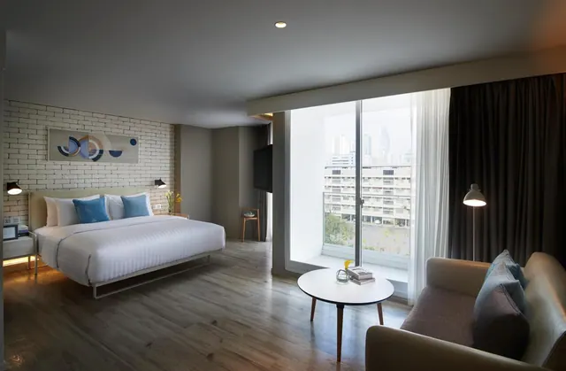 A hotel bedroom with a king bed, white brick accent wall, lounge seating and a sliding glass door looking out over city buildings.