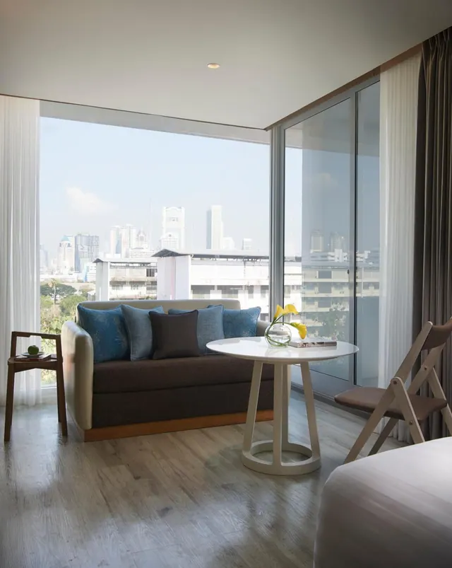 A living area with a dark grey sofa and blue cushions, a round white side table holding a vase of yellow flowers, and floor-to-ceiling windows looking out at city towers.