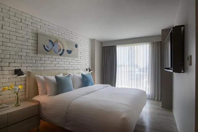 A bedroom with a made king bed, a whitewashed brick accent wall, a wall-mounted television, and a large window with sheer curtains showing tall buildings outside.