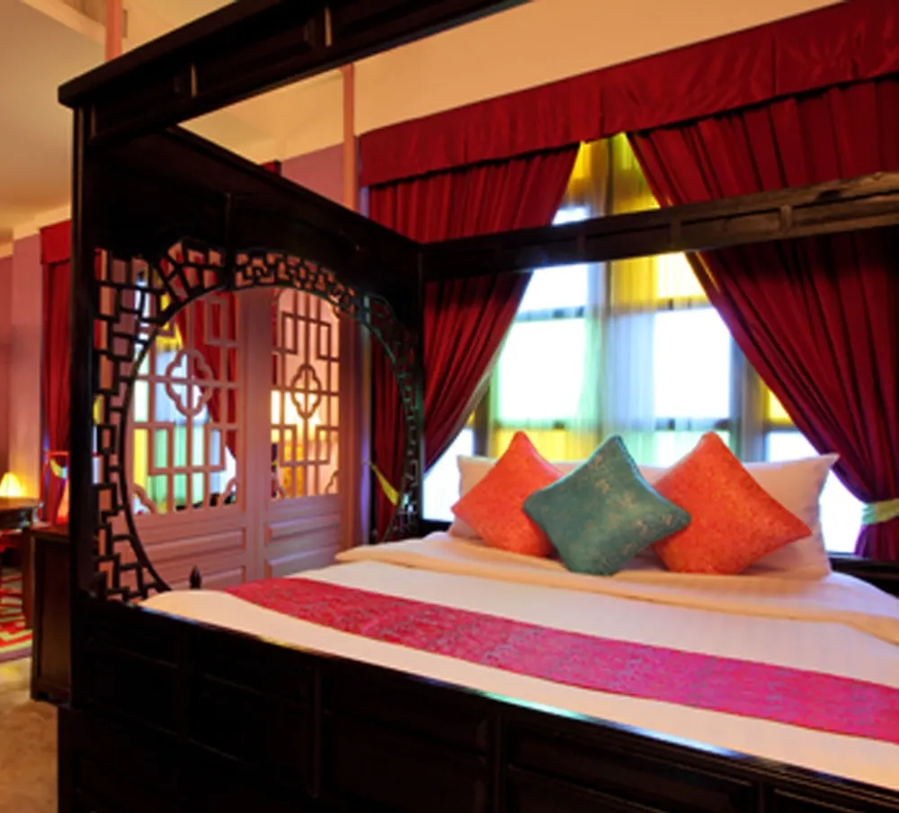 A carved dark wood four-poster bed with orange and teal cushions sits beside red curtains and a window, with a lattice-screened door to the left.