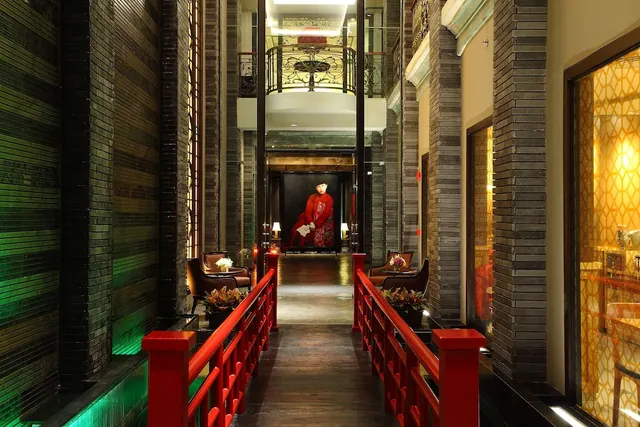 A walkway with red railings runs between dark brick columns toward a lounge with leather armchairs and a large painting of a figure in red.