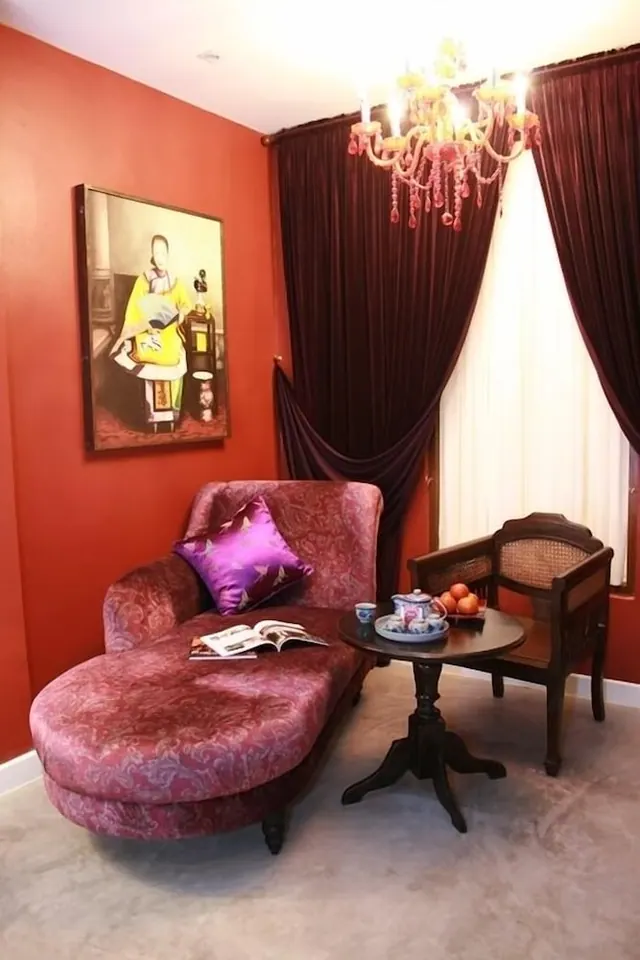 A red upholstered chaise lounge with a purple cushion and an open magazine sits beside a round wooden table holding a teapot and a tray of oranges, beneath a crystal chandelier.