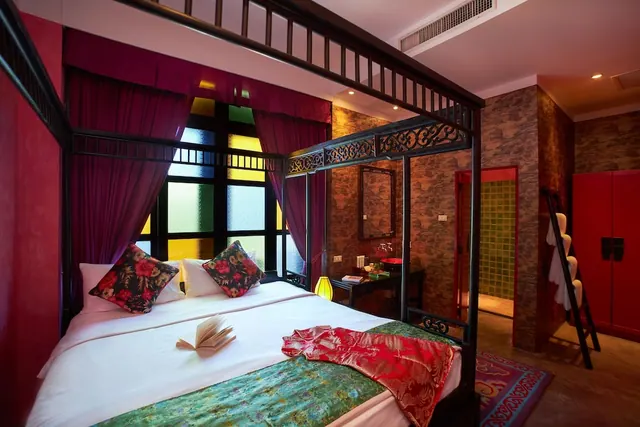 A bedroom with a canopy four-poster bed, red curtains, stained glass windows, a red wardrobe, and a doorway into a green-tiled bathroom.