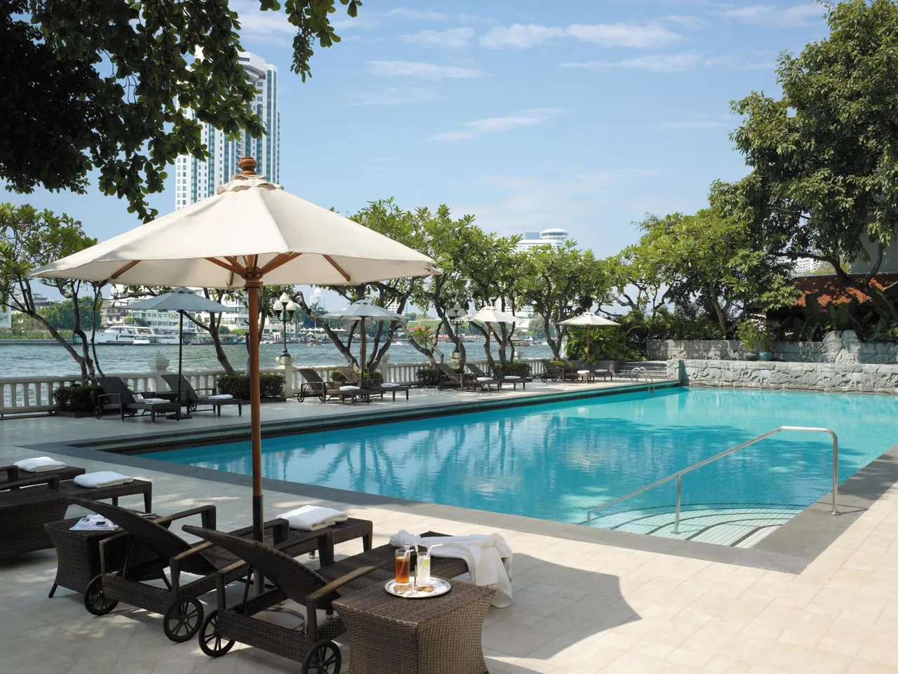 A large swimming pool with wicker sun loungers and parasol umbrellas on a tiled deck, with a river and a high-rise tower visible beyond the trees.