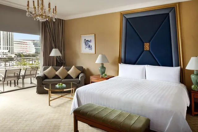 A bedroom with a king bed, a grey sofa and a round glass coffee table, with a glass door opening onto a balcony that overlooks city buildings.