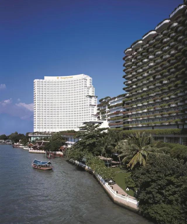 A white hotel tower and a balconied riverside wing rise above the Chao Phraya river, with a passenger boat on the water in the foreground.