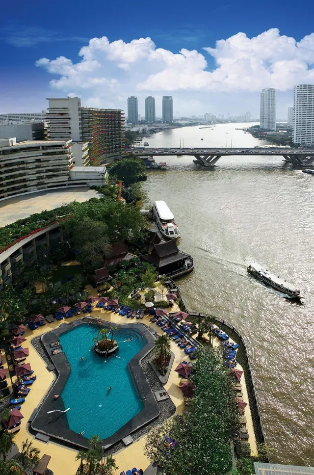 Aerial view of a hotel pool with sun loungers and umbrellas beside a wide river with boats, a bridge and city towers in the background.