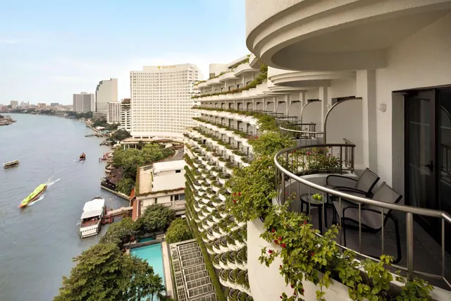 A balcony with chairs and a small table overlooks the Chao Phraya river, boats, a hotel pool and tall city towers.