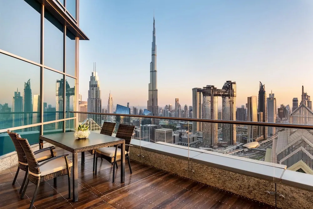 A furnished balcony with a wicker dining table and chairs overlooks a city skyline with the Burj Khalifa at dusk.