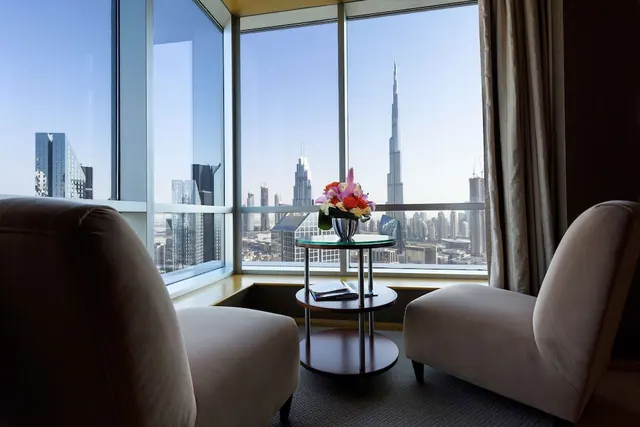 Two upholstered armchairs and a round side table with a flower arrangement face floor-to-ceiling windows showing the Burj Khalifa and a city skyline.