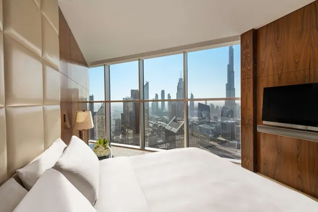 A king bed faces floor-to-ceiling windows with a city skyline and the Burj Khalifa tower in view, and a wall-mounted television is on the wood-panelled wall.