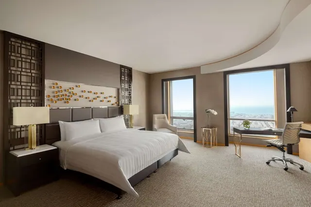 A staged hotel bedroom with a king bed, an armchair and a desk, with tall windows looking out over a city and the sea.