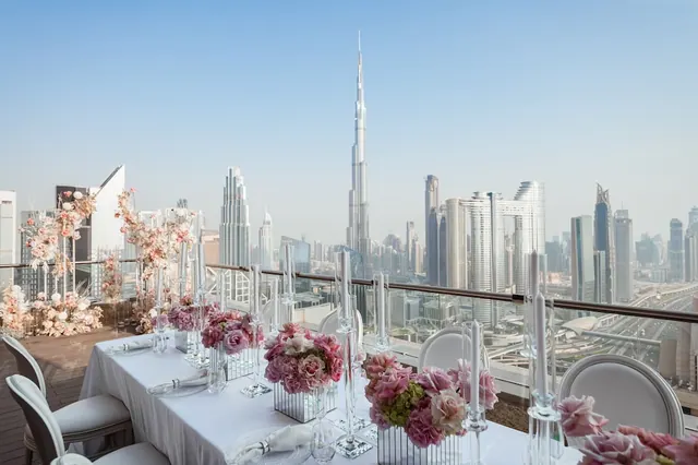 A long dining table set with white linen, pink flowers and candles on a rooftop terrace, with the Burj Khalifa and the city skyline behind it.