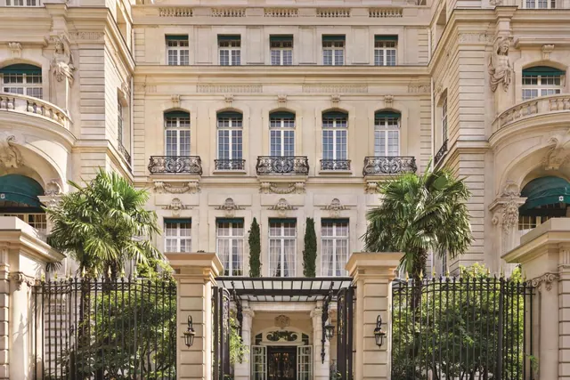 A grand historic building facade with ornate balconies, sculptures, palms, and a gated entrance.