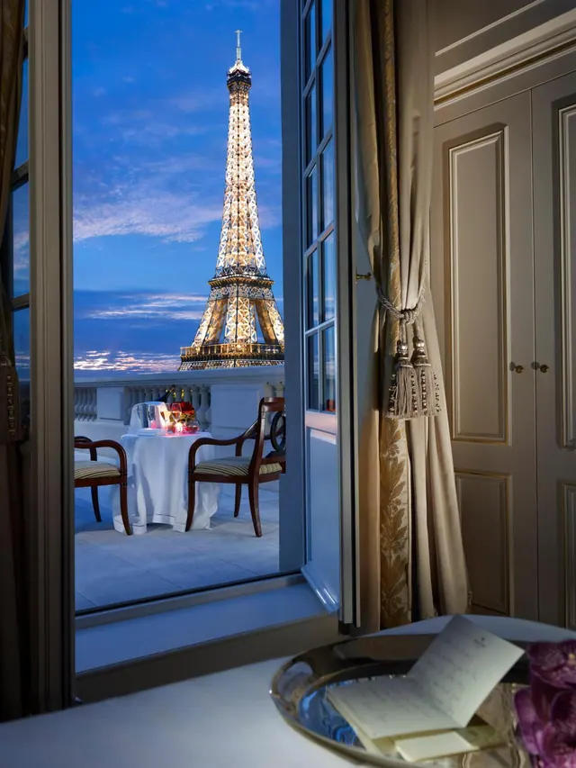 An open window looks onto a furnished terrace with the Eiffel Tower at dusk.